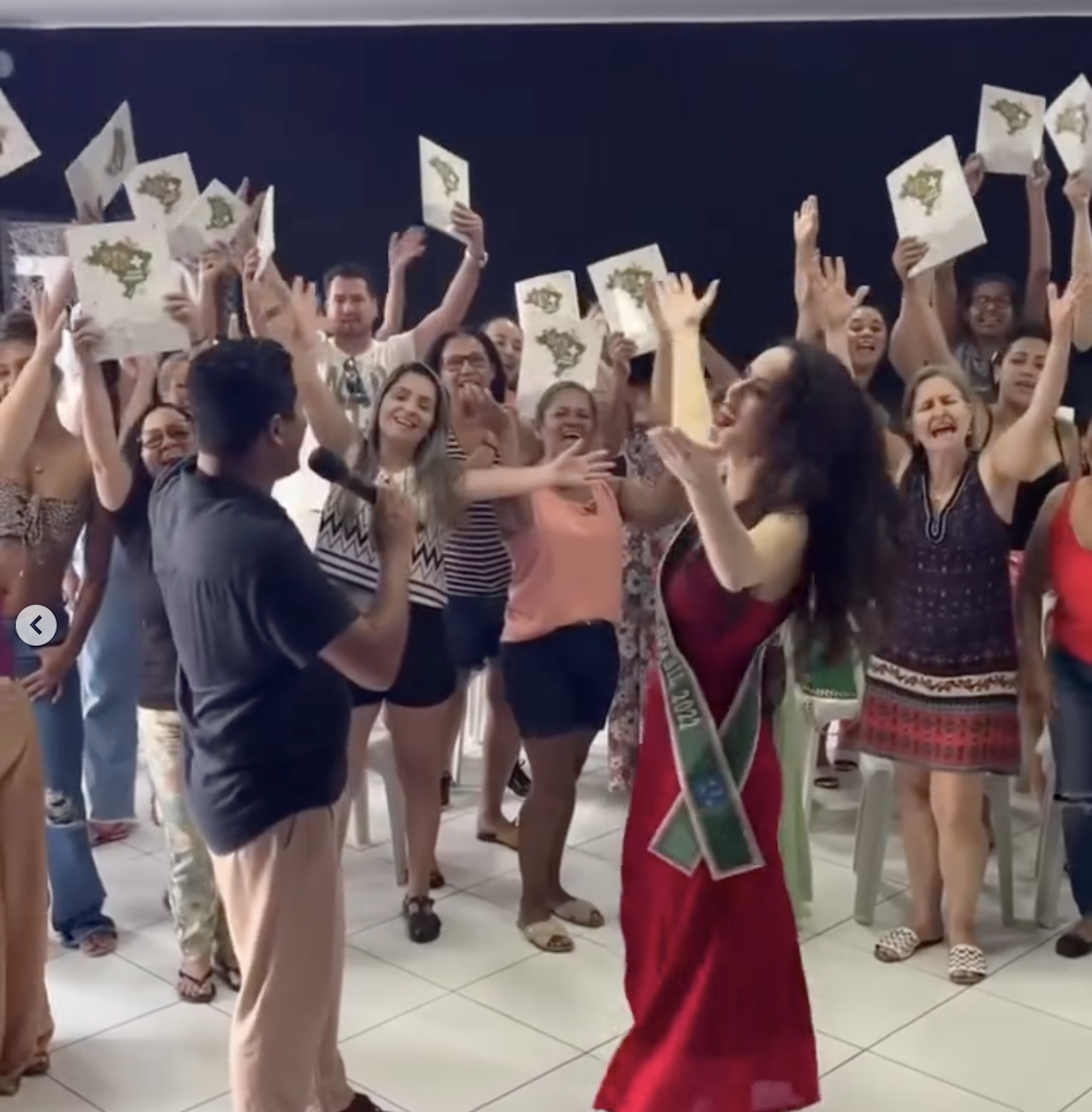 Mulher de vestido vermelho e faixa verde dança ao lado de um homem com microfone, enquanto o público comemora com as mãos levantadas